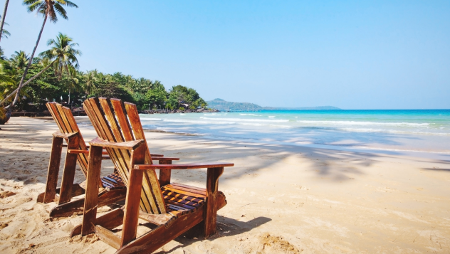 Two wooden deck chairs facing a tropical beach with clear blue sea, palm trees, and bright sunshine — perfect winter sun holiday destination.”
